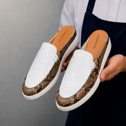 Man holding a pair of Melodiapelle Madise Mule shoes in white and snake-print brown leather, close-up