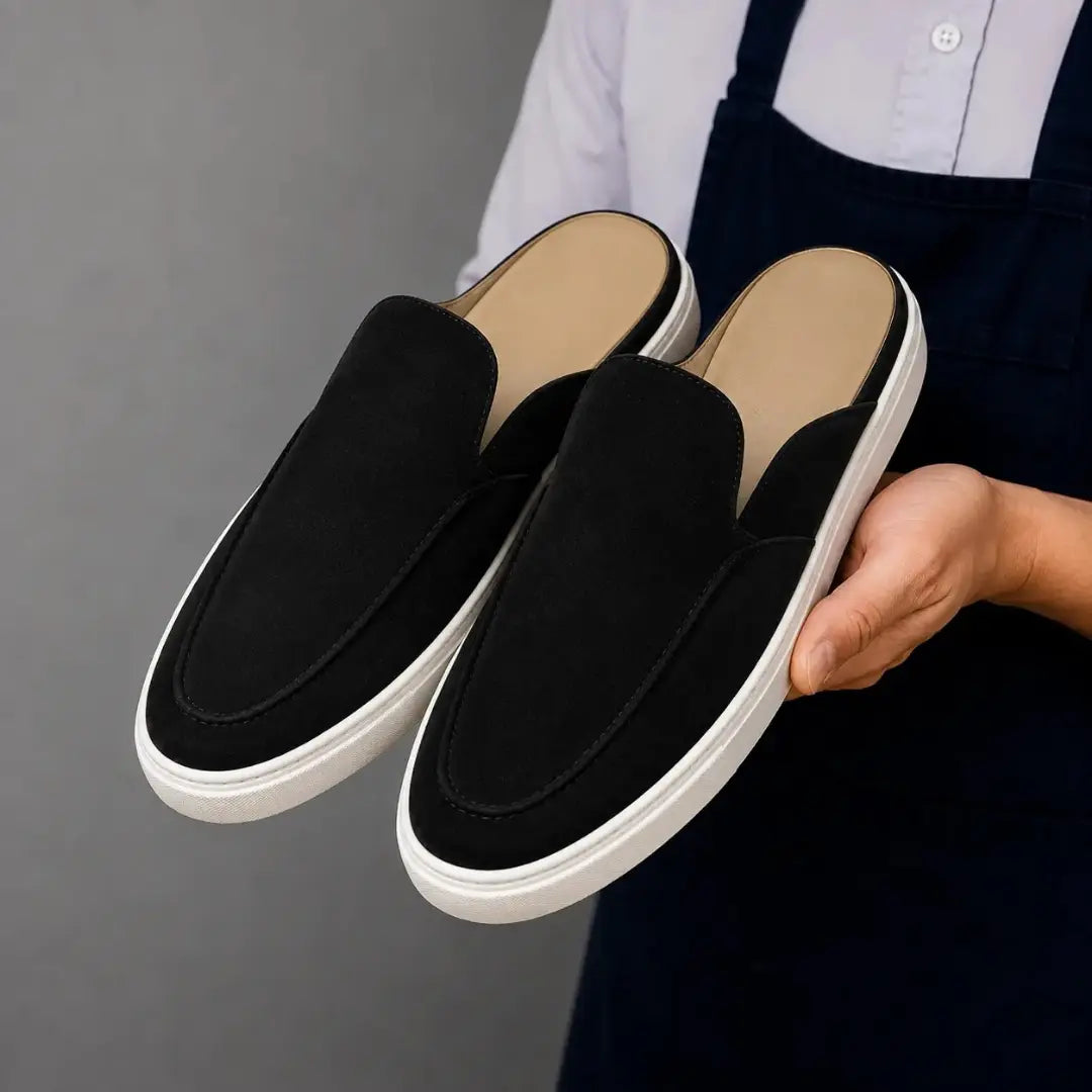 Man holding a pair of Melodiapelle Madurai Mule shoes in black suede, close-up
