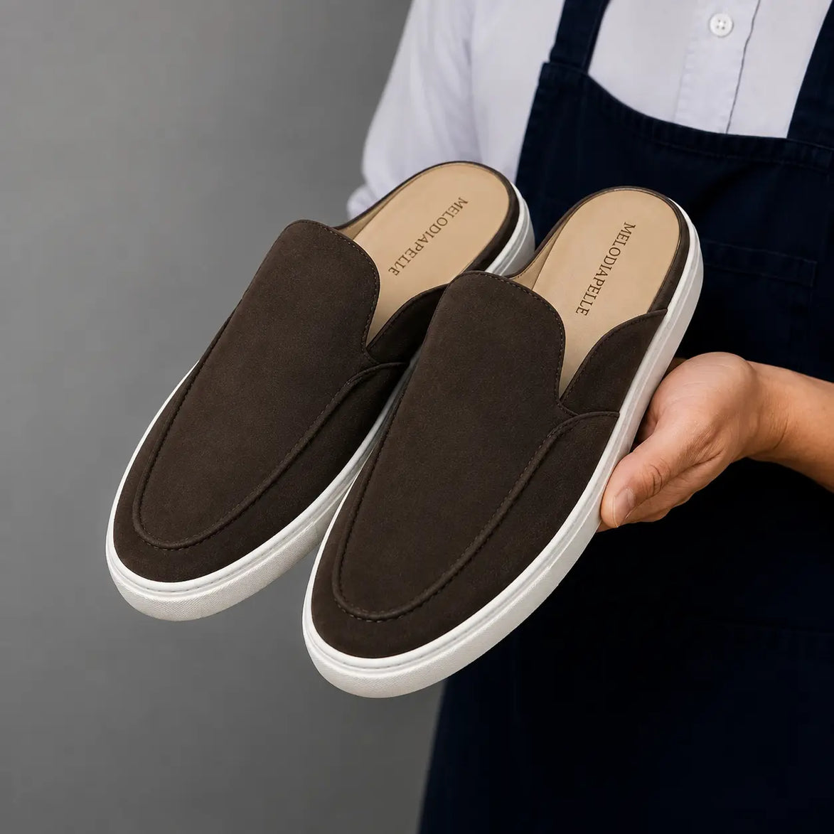 Man holding a pair of Melodiapelle Madurai Mule shoes in dark brown suede, close-up