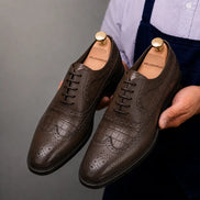 Man holding a pair of Melodiapelle Malcomb Croc-Panel Wingtip Derby shoes in brown leather, close-up