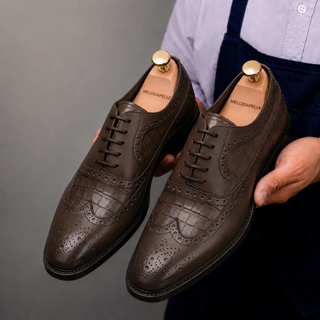 Man holding a pair of Melodiapelle Malcomb Croc-Panel Wingtip Derby shoes in brown leather, close-up