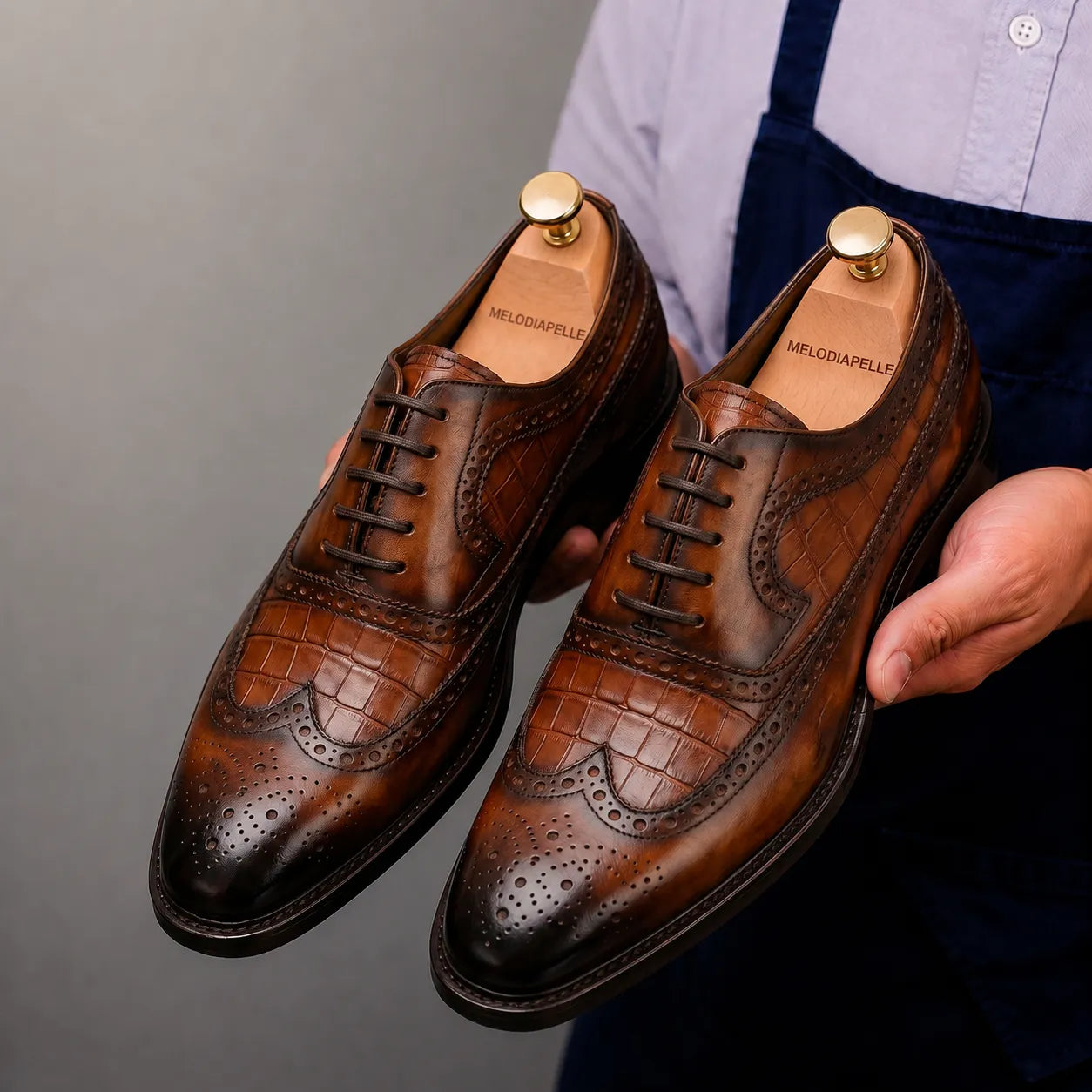 Man holding a pair of Melodiapelle Malcomb Croc-Panel Wingtip Derby shoes in cognac and dark brown leather, close-up