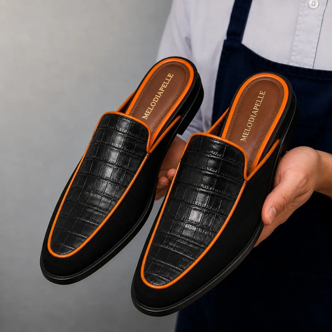 Man holding a pair of Melodiapelle Malta Mule shoes in black croc-embossed leather with orange piping, close-up
