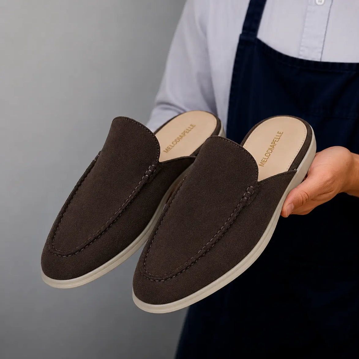 Man holding a pair of Melodiapelle Martia Mule shoes in dark brown suede, close-up