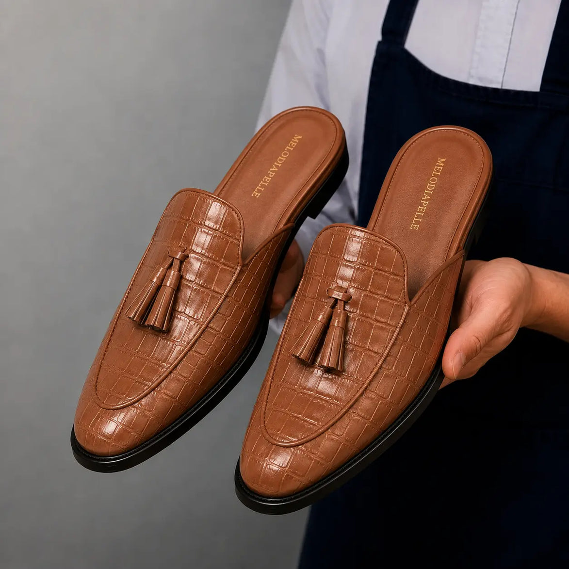Man holding a pair of Melodiapelle Mason Mule shoes in tan croc-embossed leather, close-up