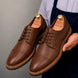 Man holding a pair of Melodiapelle Max Cap Toe Derby shoes in mole brown nubuck leather, close-up