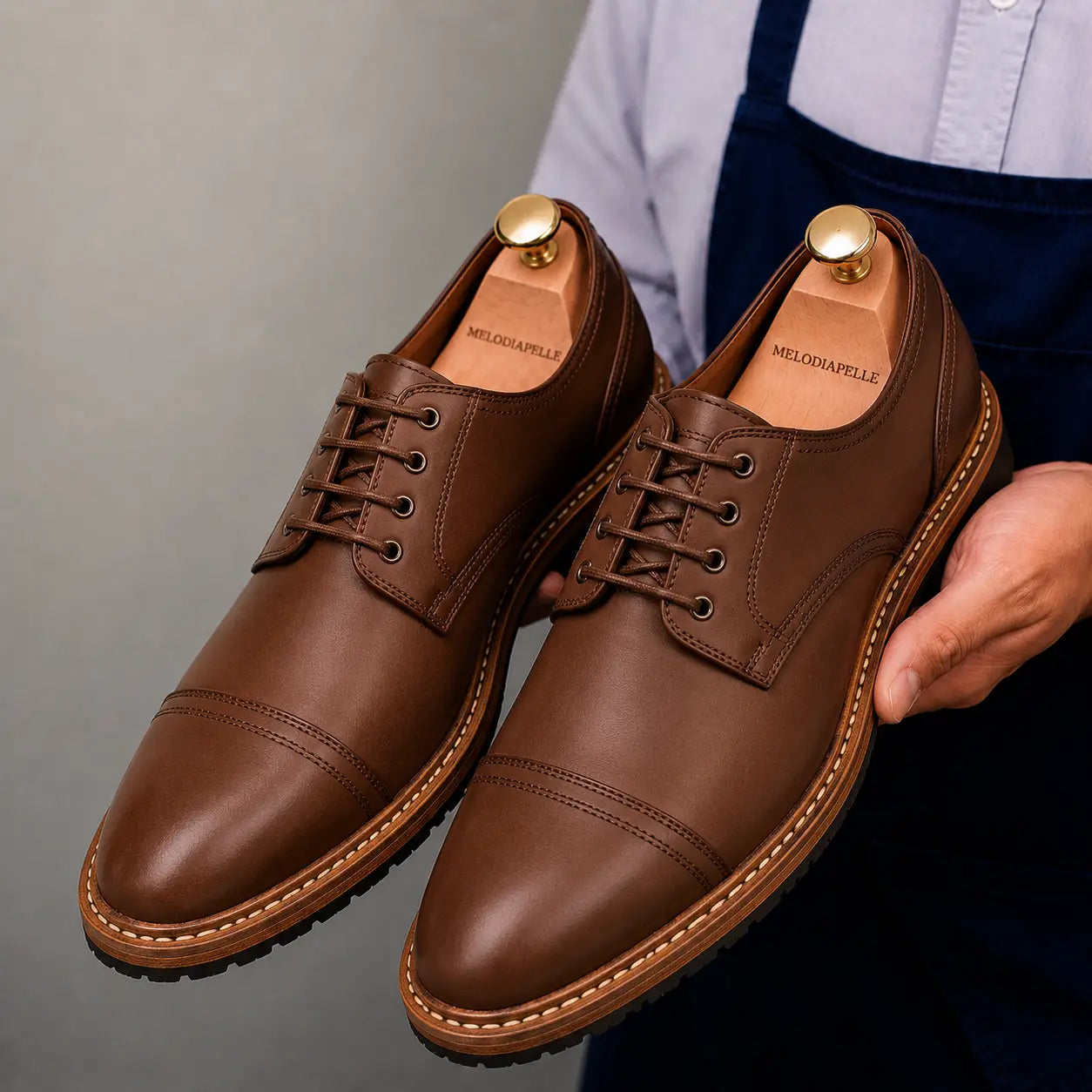 Man holding a pair of Melodiapelle Max Cap Toe Derby shoes in mole brown nubuck leather, close-up