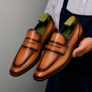 Man holding a pair of Melodiapelle Maximus Dress Penny Loafer shoes in cognac tan burnished leather, close-up