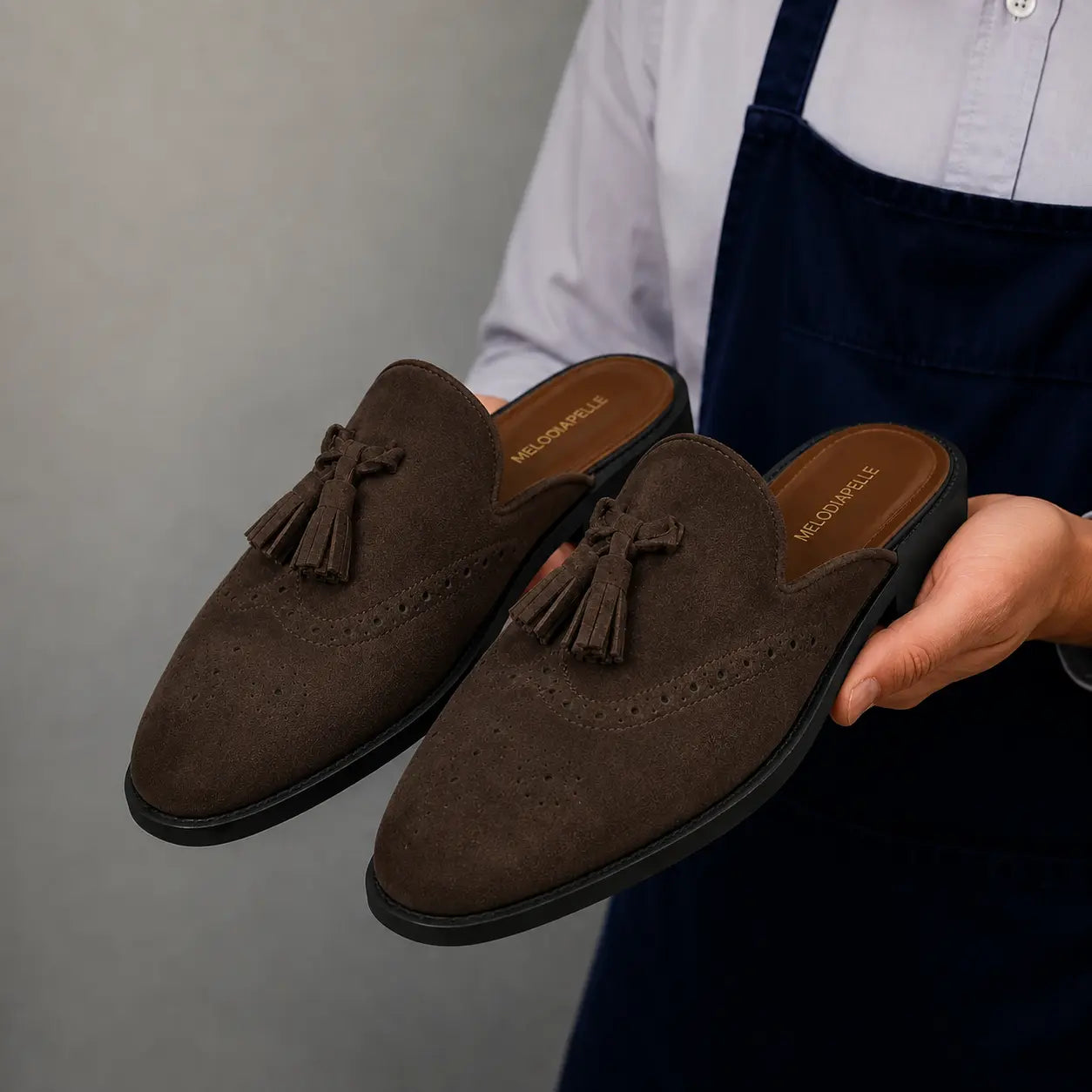 Man holding a pair of Melodiapelle Mendiai Mule shoes in dark brown suede, close-up