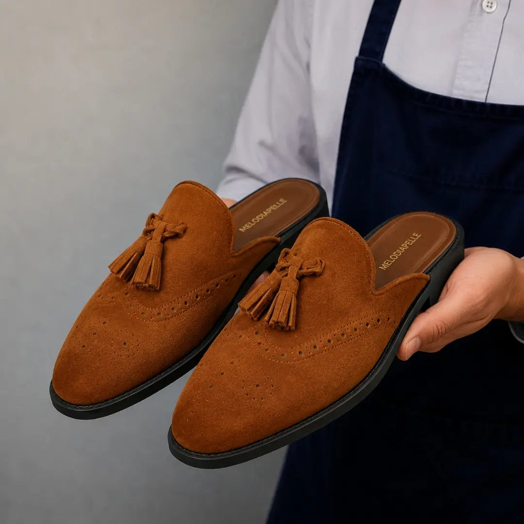 Man holding a pair of Melodiapelle Mendiai Mule shoes in rust tan suede, close-up