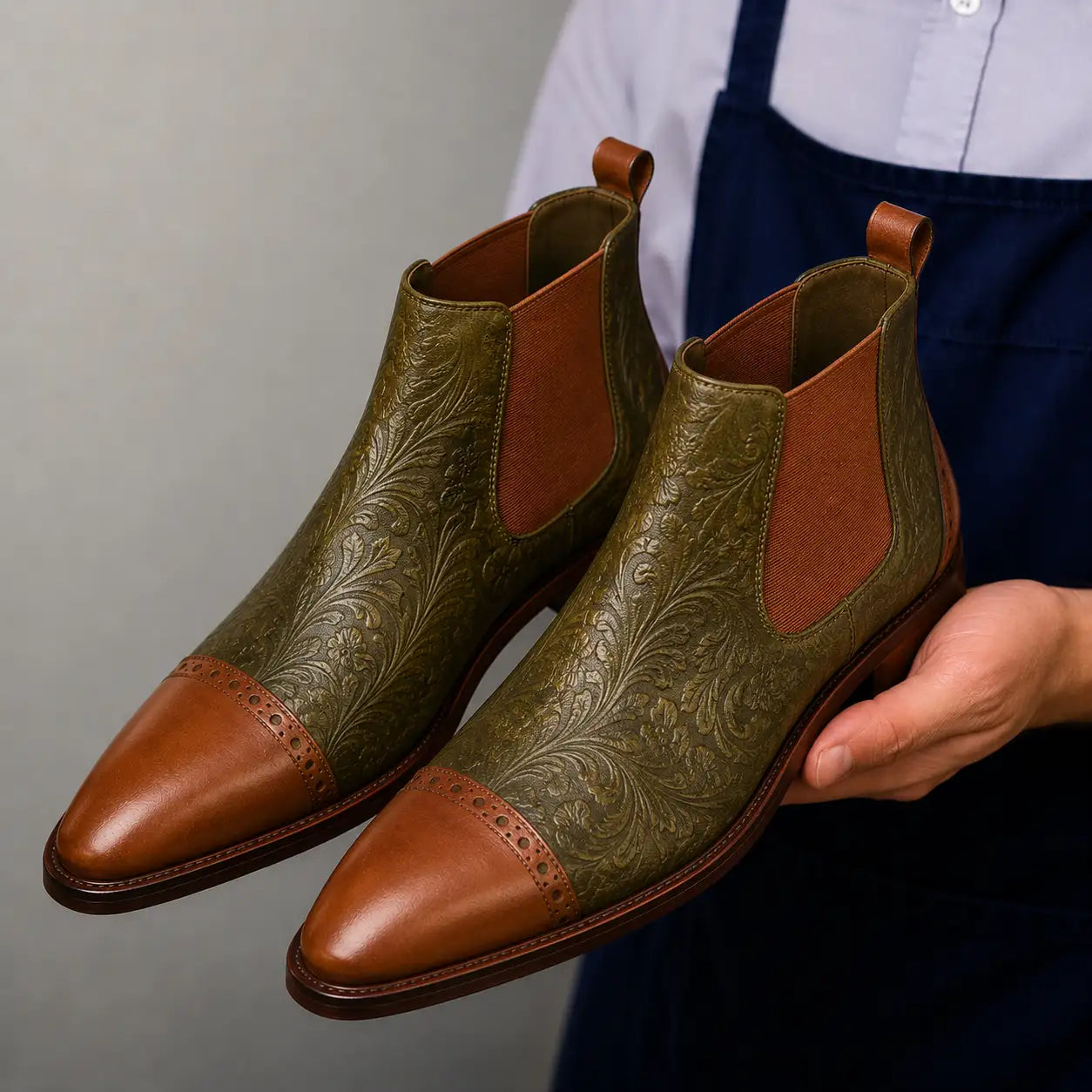 Man holding a pair of Melodiapelle Merida Floral-Embossed Chelsea Boots in olive and tan leather, close-up