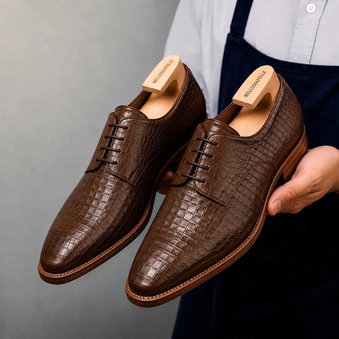 Man holding a pair of Melodiapelle Meta Wholecut Oxford shoes in taupe brown croc-embossed leather, close-up
