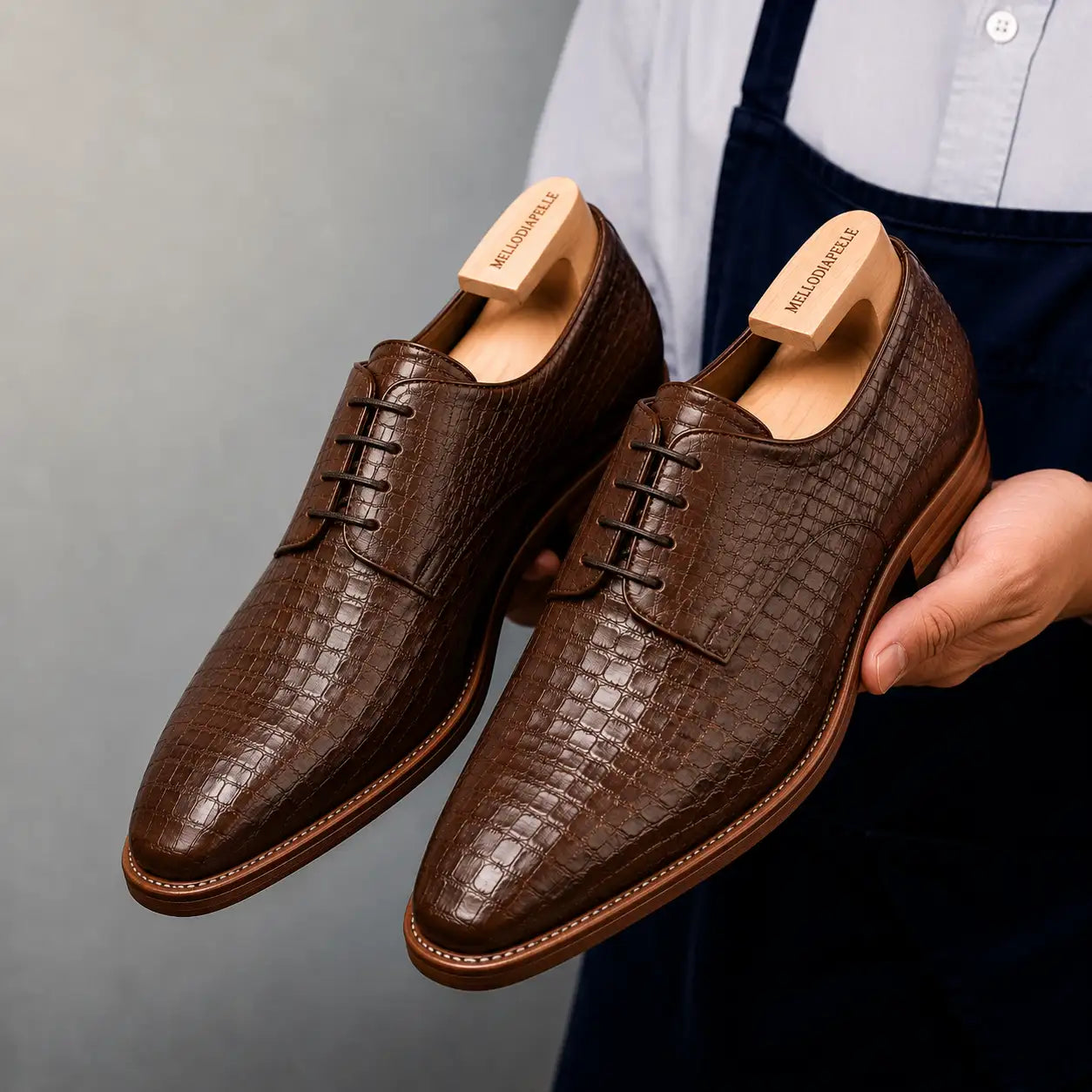Man holding a pair of Melodiapelle Meta Wholecut Oxford shoes in taupe brown croc-embossed leather, close-up