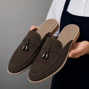 Man holding a pair of Melodiapelle Millo Mule shoes in dark brown suede, close-up