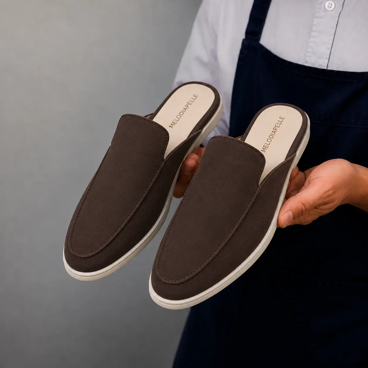 Man holding a pair of Melodiapelle Mole Mule shoes in dark brown suede, close-up