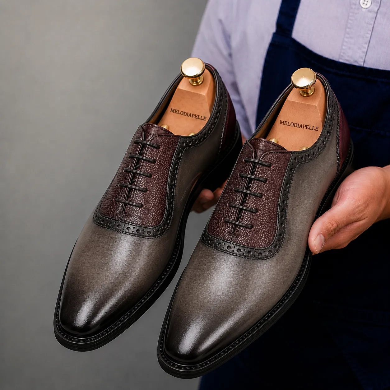 Man holding a pair of Melodiapelle Mondrian Two-Tone Saddle Oxford shoes in storm grey and bordeaux leather, close-up