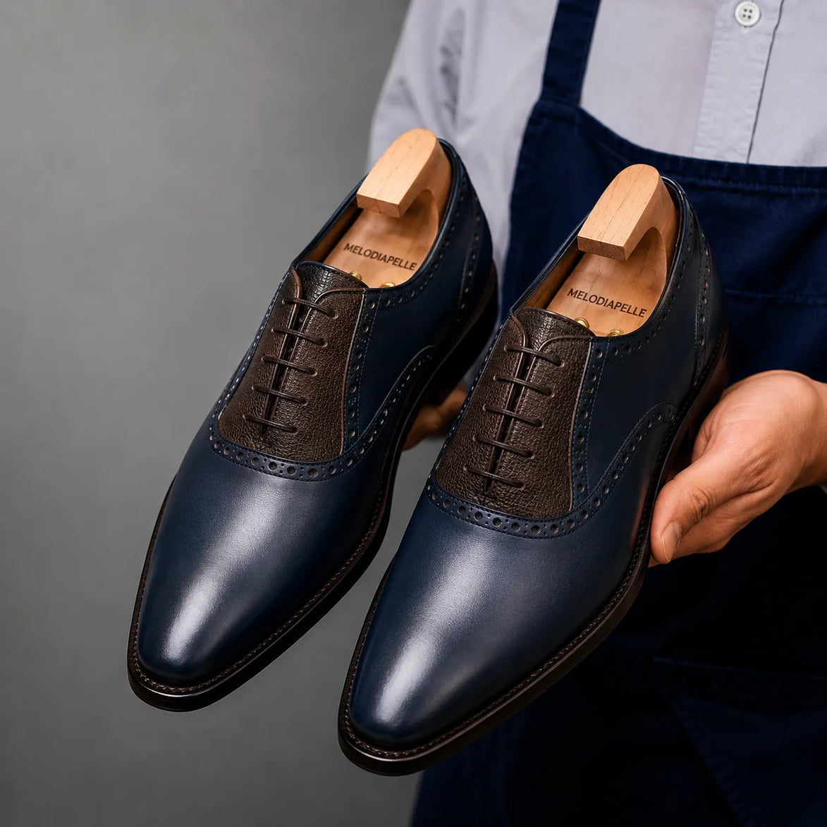 Man holding a pair of Melodiapelle Montserrat Two-Tone Saddle Oxford shoes in navy and dark brown leather, close-up