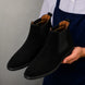 Man holding a pair of Melodiapelle Moorgate Suede Chelsea Boots in jet black suede, close-up