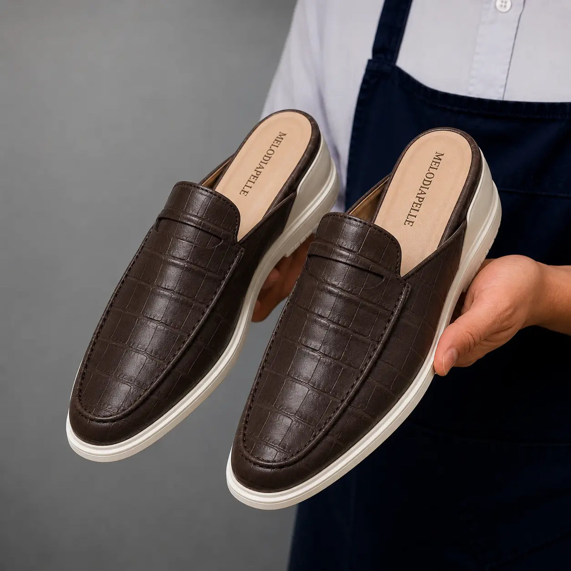 Man holding a pair of Melodiapelle Moreno Mule shoes in dark brown croc-embossed leather, close-up
