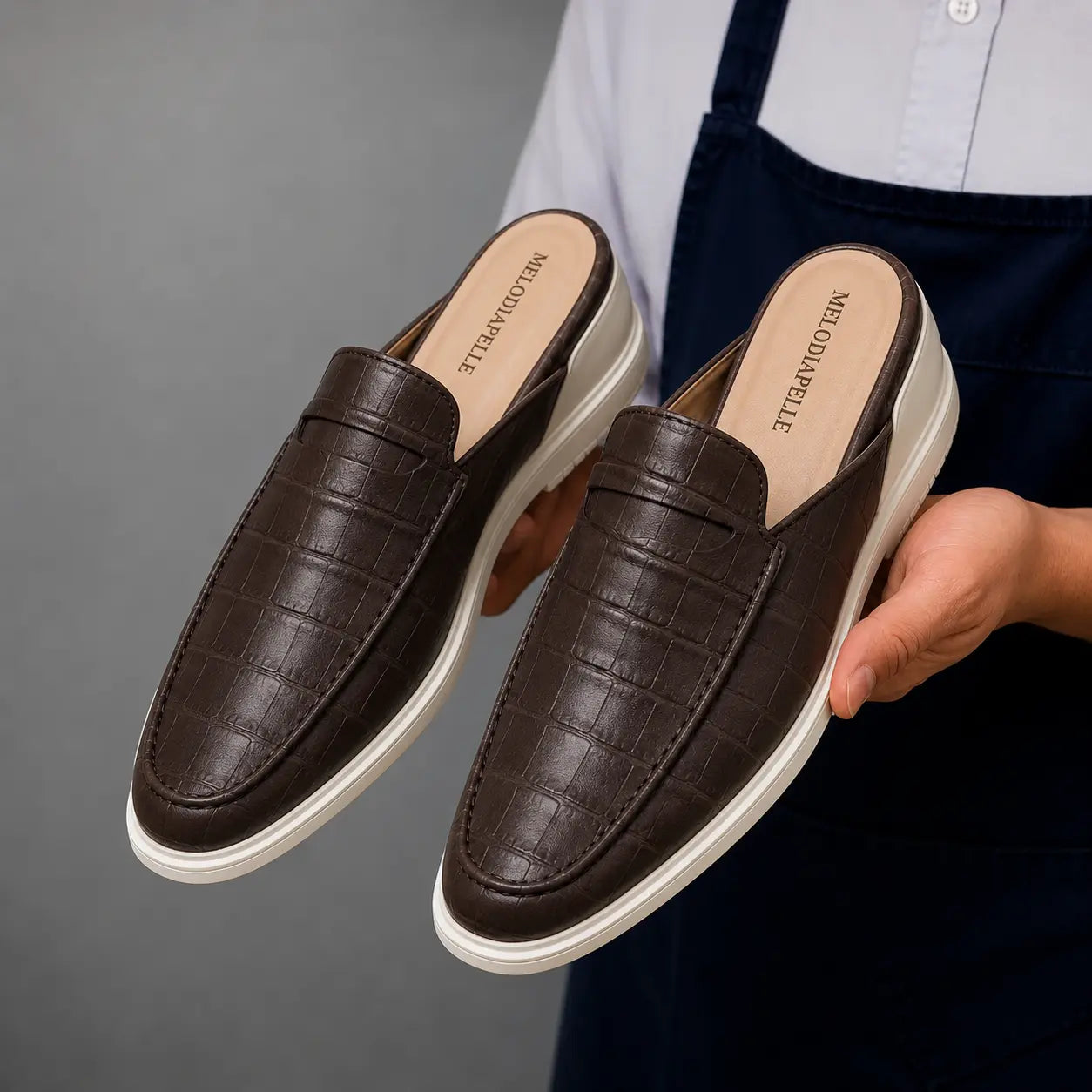 Man holding a pair of Melodiapelle Moreno Mule shoes in dark brown croc-embossed leather, close-up